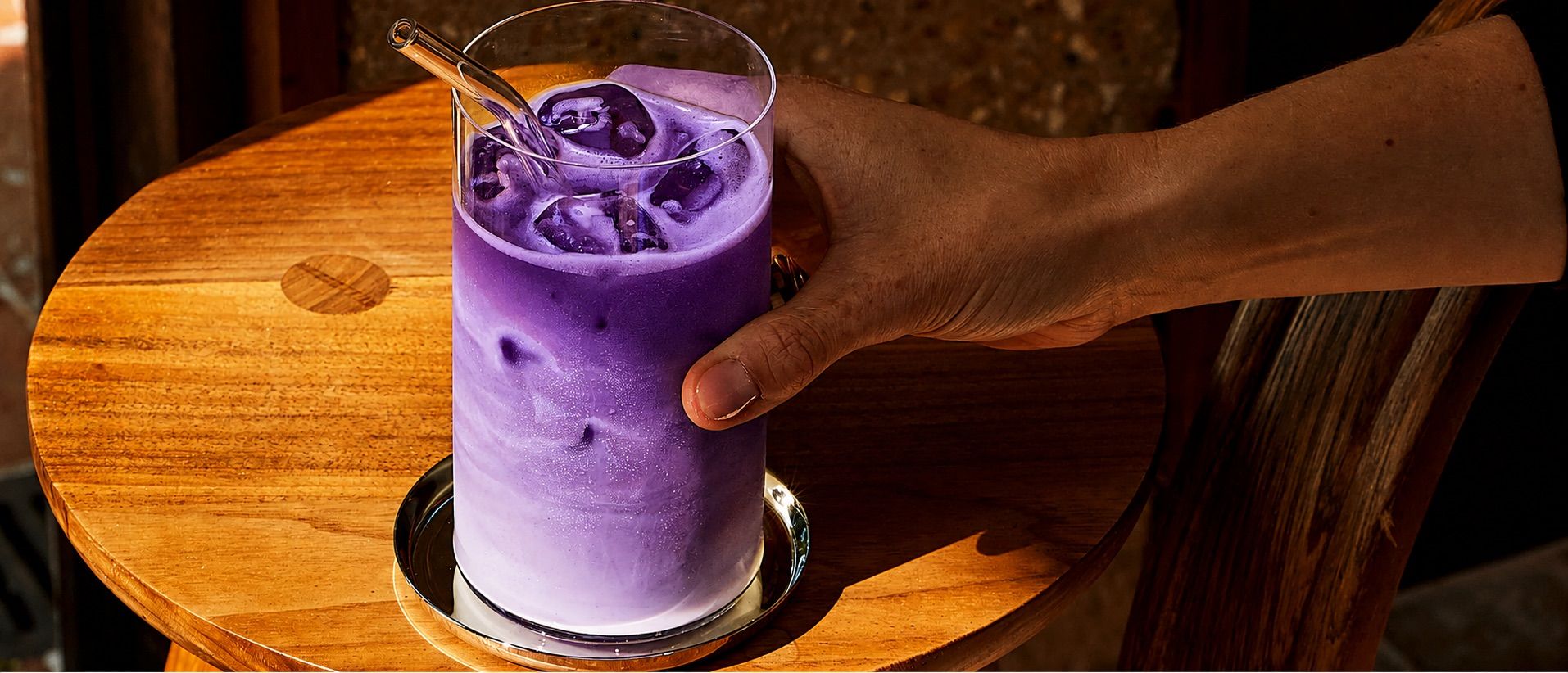 An iced ube latte in a tall glass, lilac milk over violet, set down on a wooden café table.