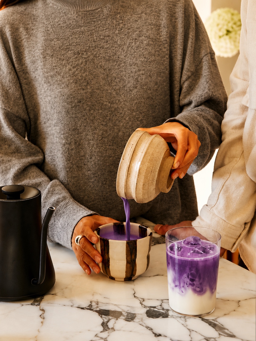 Vivid violet ube poured from a ceramic jug into a striped cup, beside an iced ube latte on a marble kitchen counter.