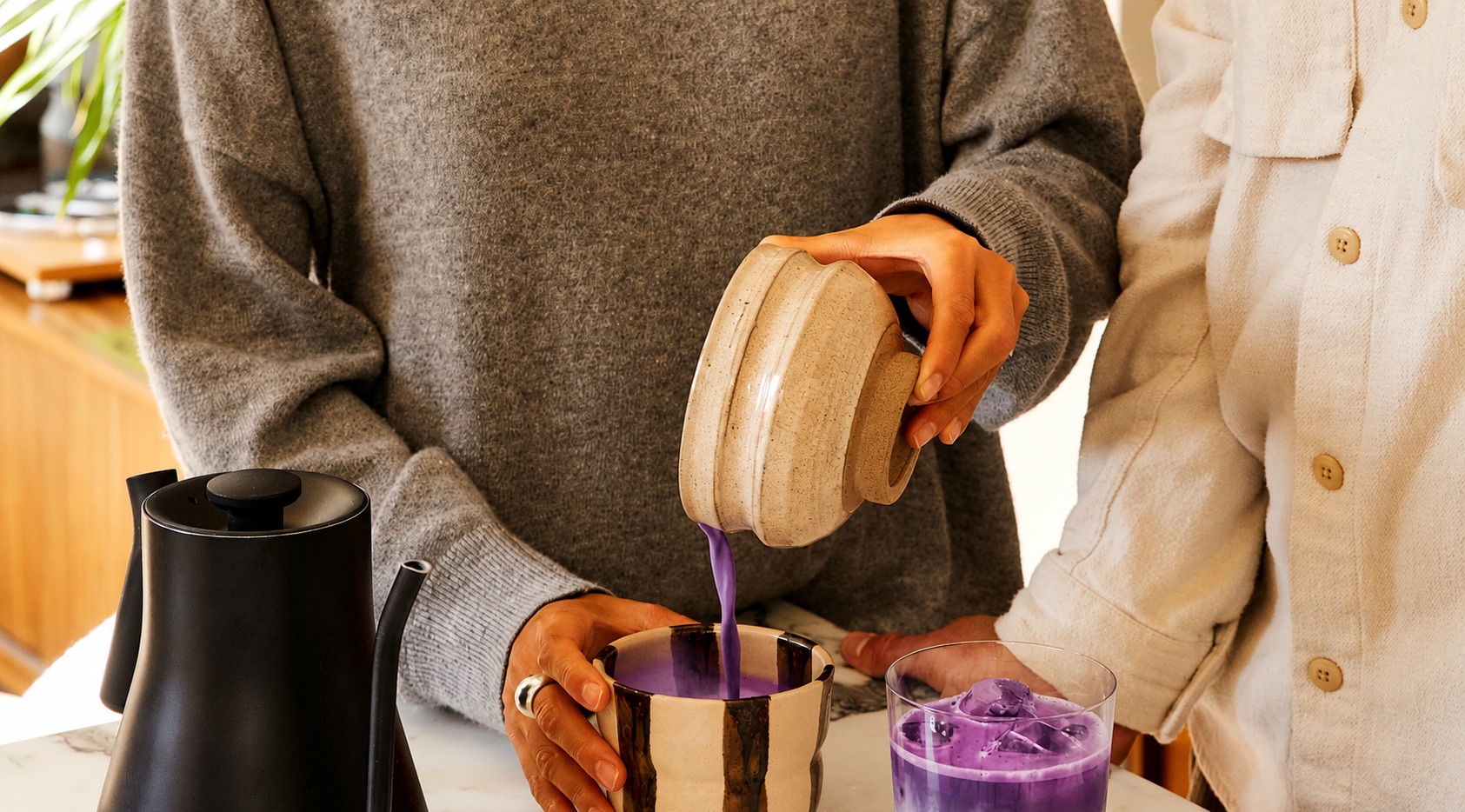 Vivid violet ube poured from a ceramic jug into a cup, beside an iced ube drink, on a marble kitchen counter.